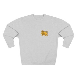 Op de afbeelding: Een lichtgrijs sweatshirt met een klein, goudkleurig embleem van een leeuw met een zwaard. De leeuw bevindt zich op de linkerbovenborst van het sweatshirt. De sweatshirt heeft een ronde hals en lange mouwen.