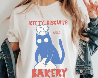May include: Cream-colored t-shirt with a blue cat wearing a chef's hat, kneading dough. The text "KITTY BISCUITS" and "BAKERY" are in red, with additional text in a smaller font. A denim jacket is draped over the shoulders.