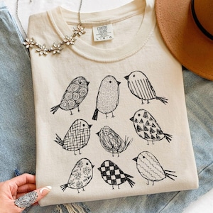 May include: Cream-colored Comfort Colors t-shirt featuring nine whimsical black bird illustrations. A brown hat, a silver necklace, and blue jeans are also in the image. The shirt has a crew neck and short sleeves.