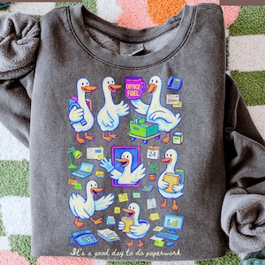 May include: Dark gray sweatshirt with a colorful graphic of cartoon geese in an office setting. The geese are depicted with office supplies and the text "It's a good day to do paperwork" is at the bottom.