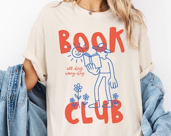 May include: Cream-colored t-shirt with a graphic design featuring the word "BOOK" in red, a blue illustration of a person reading, and the word "CLUB" in red. The text "all day every day" is also included. The shirt has short sleeves.