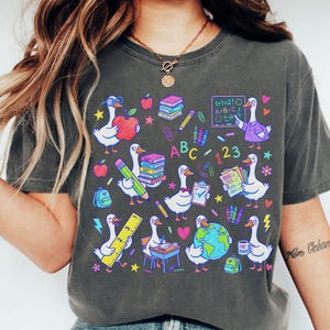 May include: Dark gray t-shirt featuring a whimsical design of cartoon geese with school supplies. The geese are depicted with books, pencils, apples, and a globe. The shirt has the text "ABC 123" and "I TEACH".