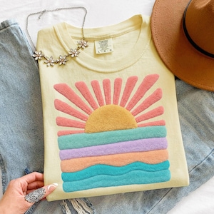 May include: A pale yellow t-shirt featuring a textured, fuzzy graphic of a sun rising over ocean waves in shades of pink, yellow, lavender, peach, and teal. The shirt has a crew neckline and a Comfort Colors label. Also includes blue jeans, a brown hat, and a silver-toned floral necklace.