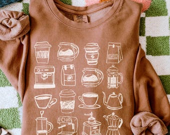 May include: A brown sweatshirt featuring a white coffee-themed graphic. The design includes various coffee-related illustrations such as cups, coffee makers, and a coffee bag. The words "FREE SHIPPING" and "ESPRESSO" are also visible.