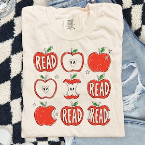 May include: A cream-colored short-sleeve t-shirt featuring a graphic print of nine red apples in various states, including whole, sliced, and eaten, with the word READ written inside several of the apple shapes, accompanied by small star icons.