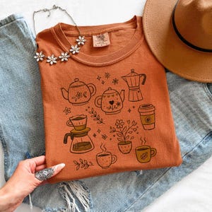 Peut inclure: Un t-shirt orange avec un th&egrave;me caf&eacute; et th&eacute;. Le t-shirt pr&eacute;sente des illustrations de th&eacute;i&egrave;res, de tasses &agrave; caf&eacute;, d'une cafeti&egrave;re et de plantes. Le t-shirt est associ&eacute; &agrave; un chapeau marron et &agrave; un jean bleu.
