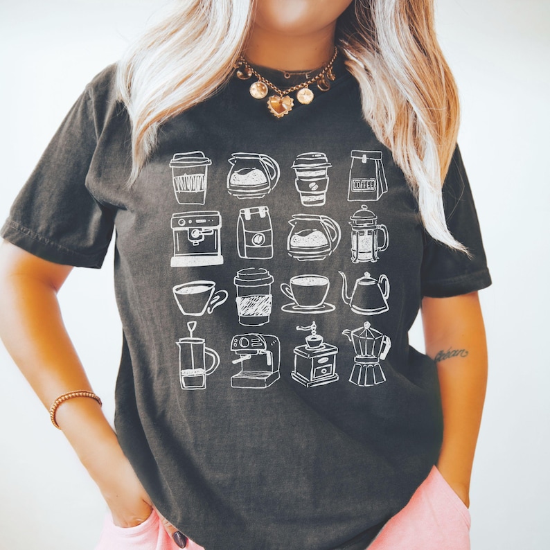 May include: Dark gray t-shirt featuring a grid of white coffee-related illustrations, including cups, coffee makers, and bags of coffee beans. The shirt has short sleeves and a relaxed fit.
