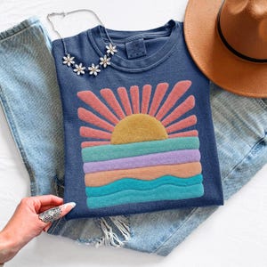 May include: A navy blue t-shirt featuring a textured, raised puff print graphic of a yellow sun rising over horizontal bands of pink, lavender, peach, and teal waves. The shirt has a crew neckline and a Comfort Colors brand label at the inner collar.