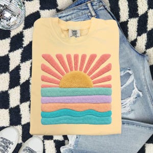 May include: A pale yellow crewneck t-shirt featuring a textured, fuzzy chenille-style graphic of a rising sun over ocean waves. The sun is golden yellow with coral rays, while the water consists of horizontal bands in teal, lavender, and peach.