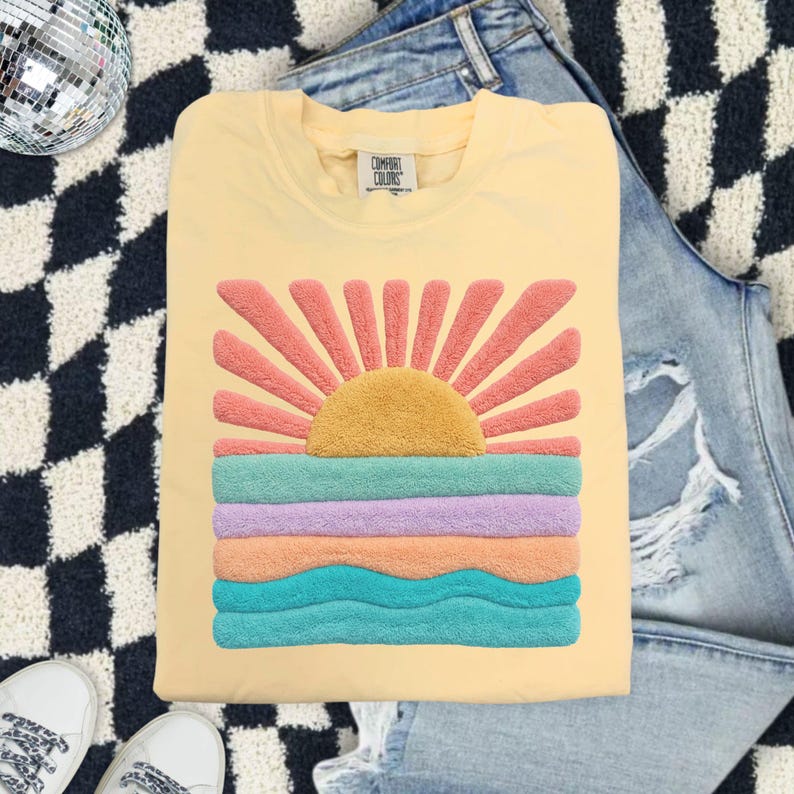 May include: A pale yellow crewneck t-shirt featuring a textured, fuzzy chenille-style graphic of a rising sun over ocean waves. The sun is golden yellow with coral rays, while the water consists of horizontal bands in teal, lavender, and peach.
