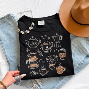Peut inclure: T-shirt noir avec des illustrations sur le th&egrave;me du caf&eacute; en blanc et marron. Le motif comprend des th&eacute;i&egrave;res, des tasses &agrave; caf&eacute; et une cafeti&egrave;re, avec des motifs floraux et en forme de c&oelig;ur. Le t-shirt est pos&eacute; sur un jean, avec un chapeau marron.