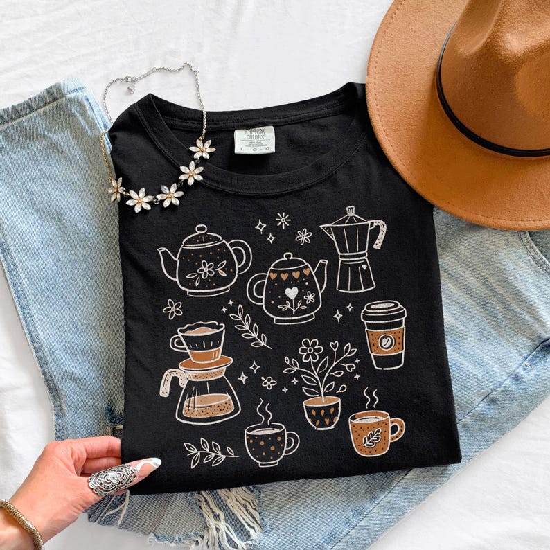 Peut inclure: T-shirt noir avec des illustrations sur le th&egrave;me du caf&eacute; en blanc et marron. Le motif comprend des th&eacute;i&egrave;res, des tasses &agrave; caf&eacute; et une cafeti&egrave;re, avec des motifs floraux et en forme de c&oelig;ur. Le t-shirt est pos&eacute; sur un jean, avec un chapeau marron.