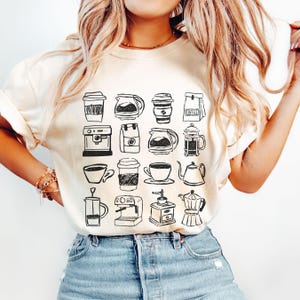 May include: Cream-colored t-shirt featuring a black and white sketch of various coffee-related items. The design includes coffee cups, coffee makers, coffee pots, and a coffee grinder. The shirt is paired with blue jeans.
