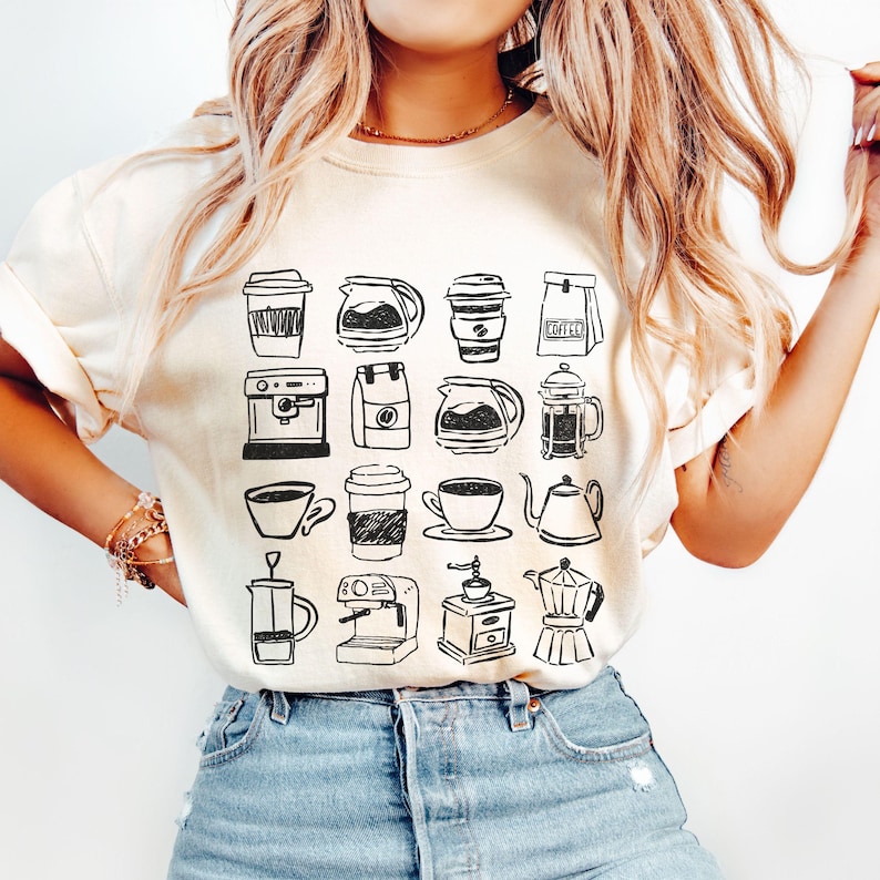 May include: Cream-colored t-shirt featuring a black and white sketch of various coffee-related items. The design includes coffee cups, coffee makers, coffee pots, and a coffee grinder. The shirt is paired with blue jeans.