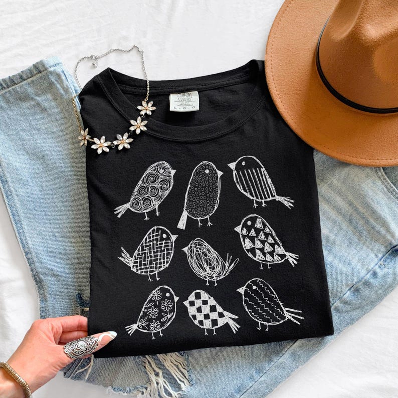 May include: Black t-shirt featuring a white illustration of nine stylized birds with various patterns. The shirt is displayed with a brown hat, a silver necklace with flower accents, and blue jeans.