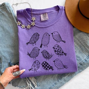 May include: A lavender Comfort Colors t-shirt featuring a black illustration of nine birds with various patterns. The shirt is paired with a floral necklace, a brown hat, and denim jeans. The Comfort Colors logo and size are visible on the shirt.