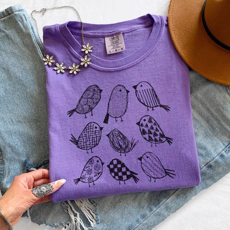 May include: A lavender Comfort Colors t-shirt featuring a black illustration of nine birds with various patterns. The shirt is paired with a floral necklace, a brown hat, and denim jeans. The Comfort Colors logo and size are visible on the shirt.
