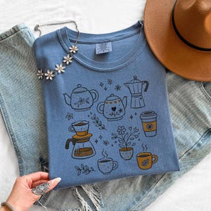 Peut inclure: T-shirt bleu clair orn&eacute; d'illustrations de th&eacute;i&egrave;res, tasses &agrave; caf&eacute; et &eacute;l&eacute;ments floraux. Le design comprend une cafeti&egrave;re. Le t-shirt est pr&eacute;sent&eacute; avec un jean bleu et un chapeau marron.