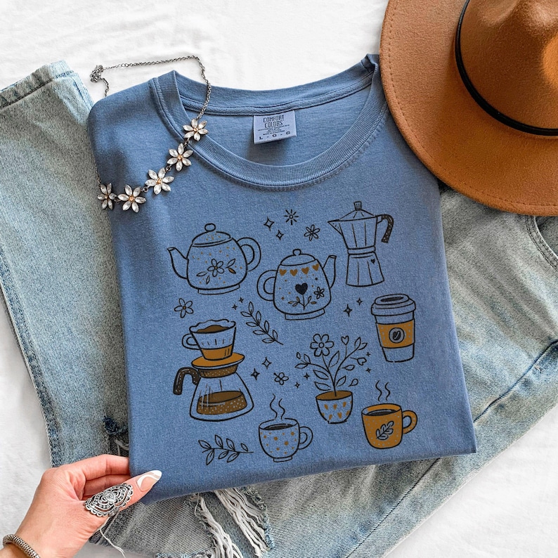 Peut inclure: T-shirt bleu clair orn&eacute; d'illustrations de th&eacute;i&egrave;res, tasses &agrave; caf&eacute; et &eacute;l&eacute;ments floraux. Le design comprend une cafeti&egrave;re. Le t-shirt est pr&eacute;sent&eacute; avec un jean bleu et un chapeau marron.