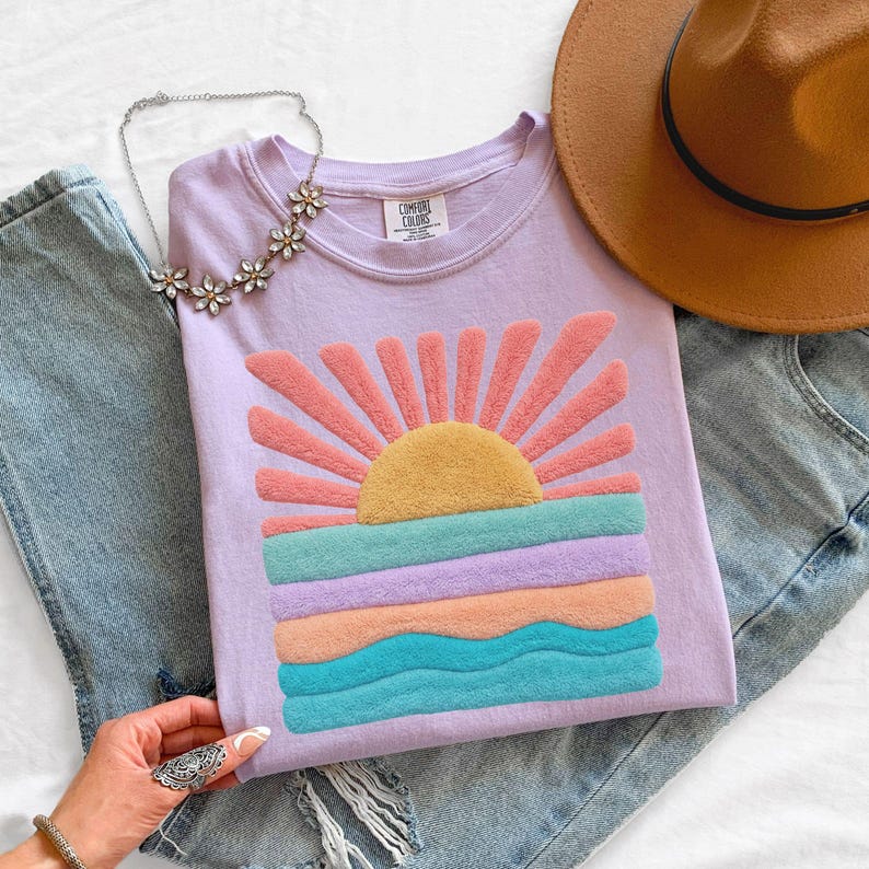 May include: A light purple t-shirt featuring a textured, raised chenille patch of a sun rising over ocean waves in shades of yellow, pink, lavender, peach, and teal. The shirt has a crew neckline with a Comfort Colors brand label inside.