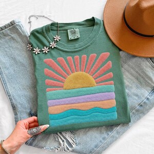 May include: A sage green short-sleeve t-shirt featuring a textured, raised graphic of a yellow sun with pink rays above horizontal wavy bands of teal, lavender, and peach. The shirt has a Comfort Colors label and is paired with blue jeans and a hat.