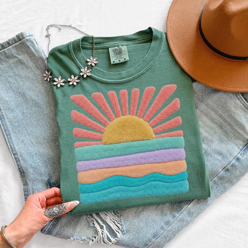 May include: A sage green short-sleeve t-shirt featuring a textured, raised graphic of a yellow sun with pink rays above horizontal wavy bands of teal, lavender, and peach. The shirt has a Comfort Colors label and is paired with blue jeans and a hat.