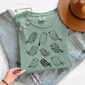 May include: Sage green t-shirt featuring a black illustration of nine stylized birds. The shirt is folded and placed on a white surface, next to a pair of blue jeans and a brown hat. A silver necklace with flower accents is also visible.