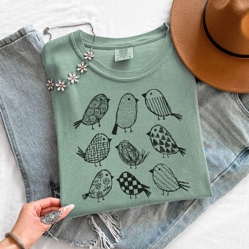 May include: Sage green t-shirt featuring a black illustration of nine stylized birds. The shirt is folded and placed on a white surface, next to a pair of blue jeans and a brown hat. A silver necklace with flower accents is also visible.