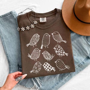 May include: A brown t-shirt featuring a white illustrated bird design. The shirt is paired with blue jeans, a brown hat, and a floral necklace. The shirt has a Comfort Colors tag.