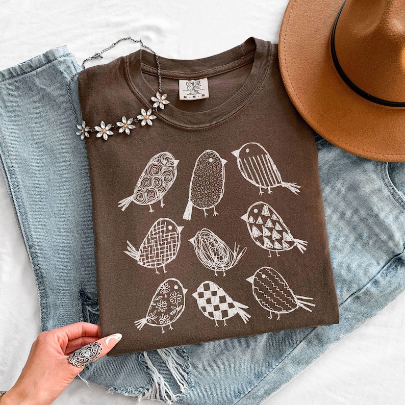 May include: A brown t-shirt featuring a white illustrated bird design. The shirt is paired with blue jeans, a brown hat, and a floral necklace. The shirt has a Comfort Colors tag.