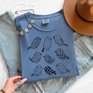 May include: A blue t-shirt with a crew neck featuring a black illustration of nine different birds. A silver necklace with flower-shaped pendants and a brown hat are also visible.