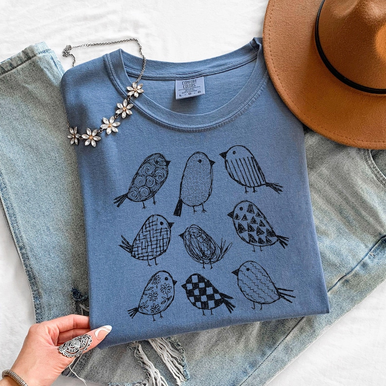 May include: A blue t-shirt with a crew neck featuring a black illustration of nine different birds. A silver necklace with flower-shaped pendants and a brown hat are also visible.