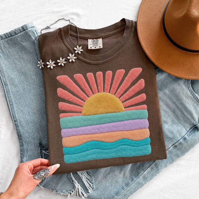 May include: A dark brown short-sleeve t-shirt featuring a textured, fuzzy graphic of a yellow sun with pink rays above horizontal bands of teal, lavender, peach, and blue waves. The shirt has a Comfort Colors label at the inner neckline.
