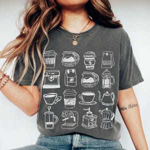 May include: Dark gray t-shirt featuring a white line drawing of various coffee-related items, including coffee cups, coffee makers, and coffee pots. The shirt is a relaxed fit and has short sleeves.