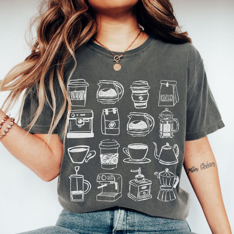 May include: Dark gray t-shirt featuring a white line drawing of various coffee-related items, including coffee cups, coffee makers, and coffee pots. The shirt is a relaxed fit and has short sleeves.
