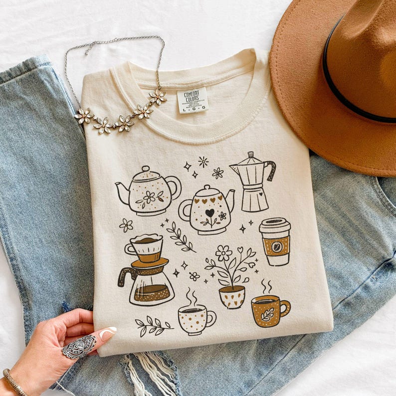 Peut inclure: Sweat-shirt cr&egrave;me avec un motif sur le th&egrave;me du caf&eacute; et du th&eacute;. Le motif comprend des th&eacute;i&egrave;res, des cafeti&egrave;res, des tasses &agrave; caf&eacute; et des &eacute;l&eacute;ments floraux. Le t-shirt est associ&eacute; &agrave; un jean bleu et un chapeau marron.