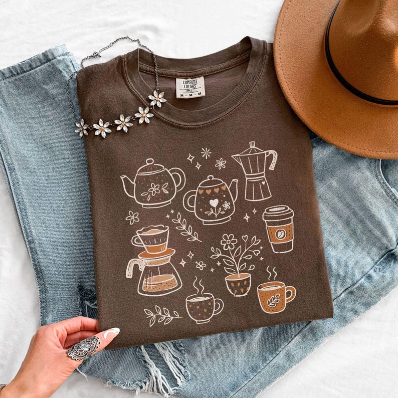 Peut inclure: Un t-shirt marron avec un motif blanc repr&eacute;sentant des &eacute;l&eacute;ments li&eacute;s au caf&eacute;, comme des th&eacute;i&egrave;res et des tasses. Le t-shirt est pr&eacute;sent&eacute; avec un jean bleu et un chapeau marron. Un collier floral est pos&eacute; sur le t-shirt.