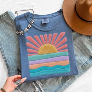 May include: A blue t-shirt featuring a textured, raised graphic of a yellow sun with pink rays above wavy bands of teal, lavender, and peach. The shirt includes a Comfort Colors label at the neck, paired with light blue denim jeans and a brown hat.