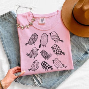 May include: A light pink t-shirt with a crew neck featuring a black illustrated bird design. The shirt is folded on top of blue denim jeans. A silver necklace with flower-shaped pendants is placed on the shirt.