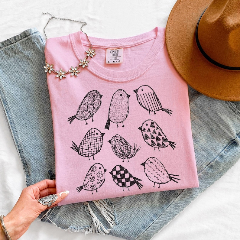 May include: A light pink t-shirt with a crew neck featuring a black illustrated bird design. The shirt is folded on top of blue denim jeans. A silver necklace with flower-shaped pendants is placed on the shirt.