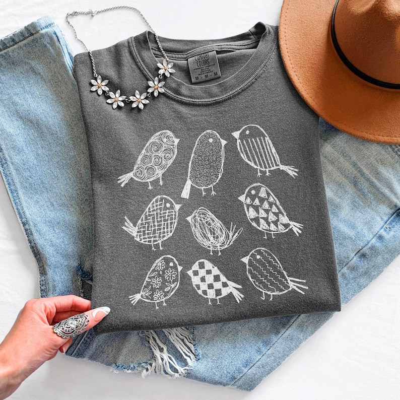 May include: A dark gray t-shirt featuring a white illustration of nine different birds. The shirt is laid on top of blue denim shorts and a pair of jeans. A silver necklace with flower-shaped pendants and a brown hat are also visible.