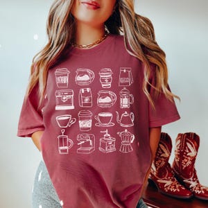 May include: A maroon t-shirt featuring a white line drawing of various coffee-related items, including coffee makers, cups, and a coffee grinder. The shirt is a short-sleeved crew neck.