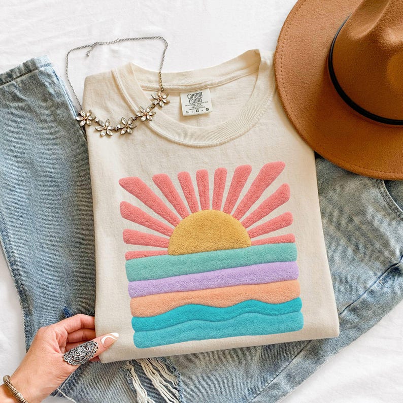 May include: A cream-colored t-shirt featuring a textured, fuzzy graphic of a rising sun over ocean waves in shades of pink, yellow, teal, lavender, and peach. The shirt is paired with blue denim jeans, a brown wide-brimmed hat, and a floral necklace.