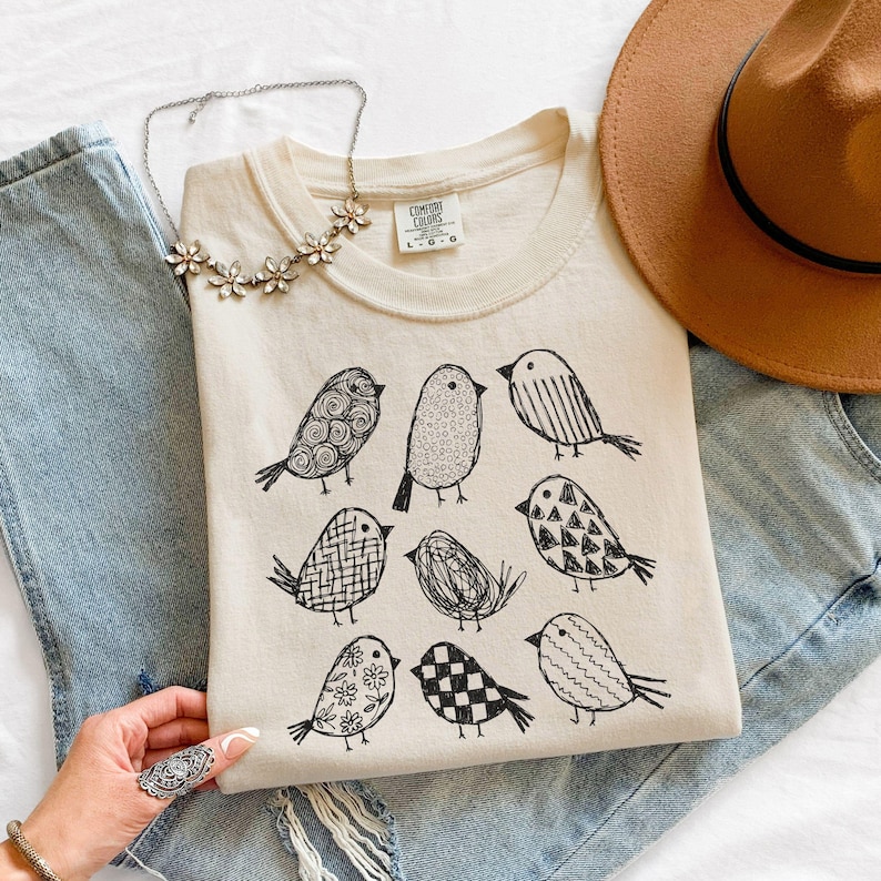 May include: Cream-colored Comfort Colors t-shirt featuring nine whimsical black bird illustrations. A brown hat, a silver necklace, and blue jeans are also in the image. The shirt has a crew neck and short sleeves.