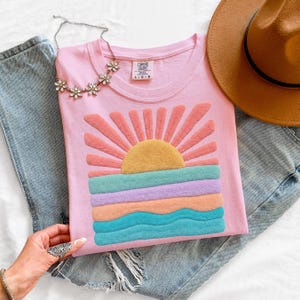 May include: A light pink t-shirt featuring a textured, raised graphic of a sun rising over ocean waves in shades of yellow, coral, purple, and teal. The shirt has a crew neckline with a Comfort Colors label and is paired with blue denim jeans.