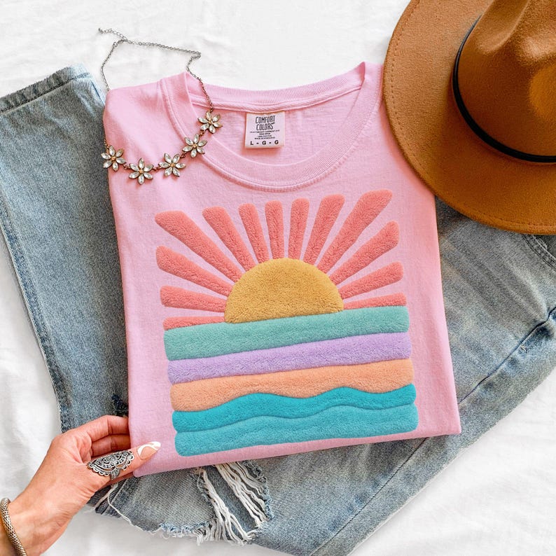 May include: A light pink t-shirt featuring a textured, raised graphic of a sun rising over ocean waves in shades of yellow, coral, purple, and teal. The shirt has a crew neckline with a Comfort Colors label and is paired with blue denim jeans.