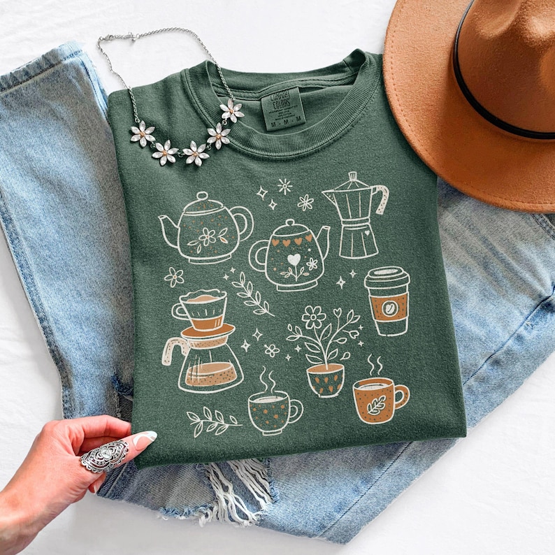 Peut inclure: T-shirt vert avec des illustrations blanches et oranges de th&eacute;i&egrave;res, tasses &agrave; caf&eacute; et fleurs. Le t-shirt est pr&eacute;sent&eacute; sur un jean bleu, avec un chapeau marron et un collier floral. Le t-shirt porte le texte "LUCKY COLORS" sur l'&eacute;tiquette.