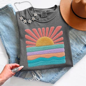 May include: A charcoal gray t-shirt featuring a textured, fuzzy graphic of a rising sun over ocean waves in shades of yellow, pink, purple, and teal. The shirt is paired with light blue denim jeans, a brown wide-brimmed hat, and a silver flower necklace.