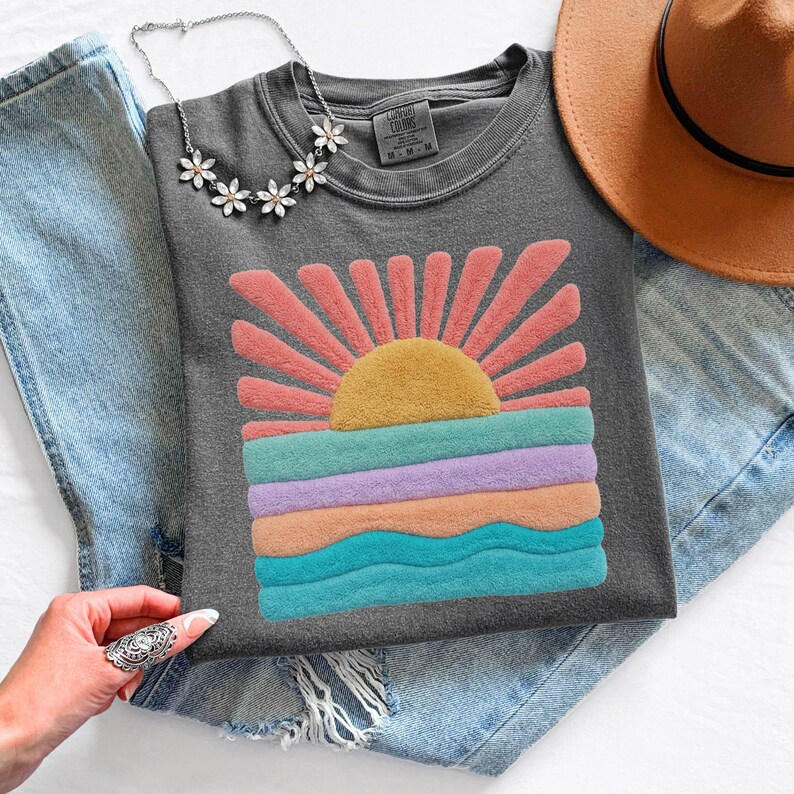 May include: A charcoal gray t-shirt featuring a textured, fuzzy graphic of a rising sun over ocean waves in shades of yellow, pink, purple, and teal. The shirt is paired with light blue denim jeans, a brown wide-brimmed hat, and a silver flower necklace.