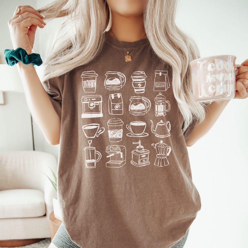 May include: A brown t-shirt featuring a white line drawing of various coffee-related items, including coffee cups, coffee makers, and coffee pots. The shirt is worn by a person holding a pink mug with the text "cool mom club".